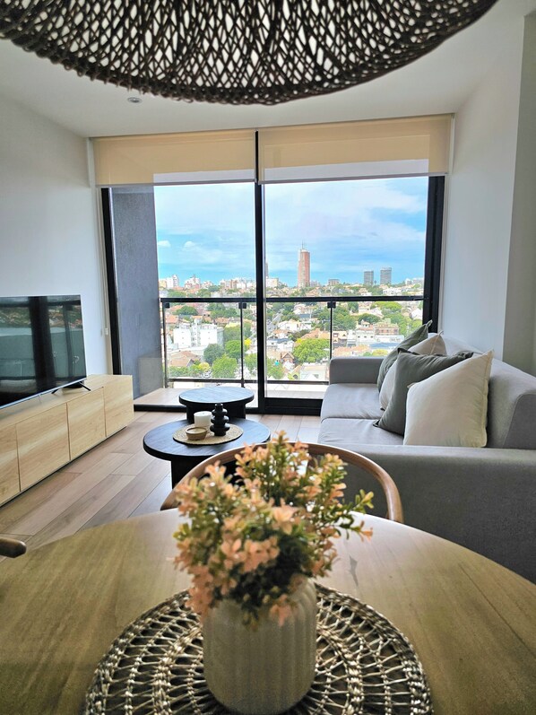 A modern living room with a sofa, a TV, a coffee table, and a large window offering a city view. - Urban 1303 | 2 rooms with garage (Mar del Plata)