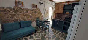 Een kamer met een stenen muur, een blauwgroene bank, een eettafel met stoelen en een keukenhoek met houten kastjes en een koelkast.