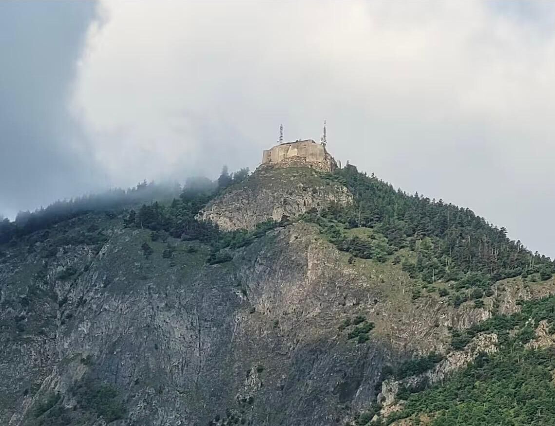 Ein Berg mit einem Gebäude auf der Spitze, umgeben von Bäumen und felsigem Gelände.