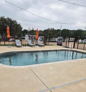 A swimming pool with lounge chairs and umbrellas, surrounded by a fence and RVs in the background.