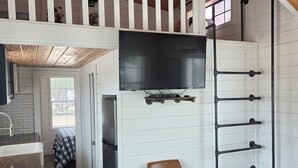Une pièce de style « loft », avec un plafond en bois, un téléviseur fixé au mur, une rambarde blanche et une fenêtre.