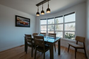 Een eetkamer met een houten tafel en stoelen, een schilderij aan de muur en grote ramen.