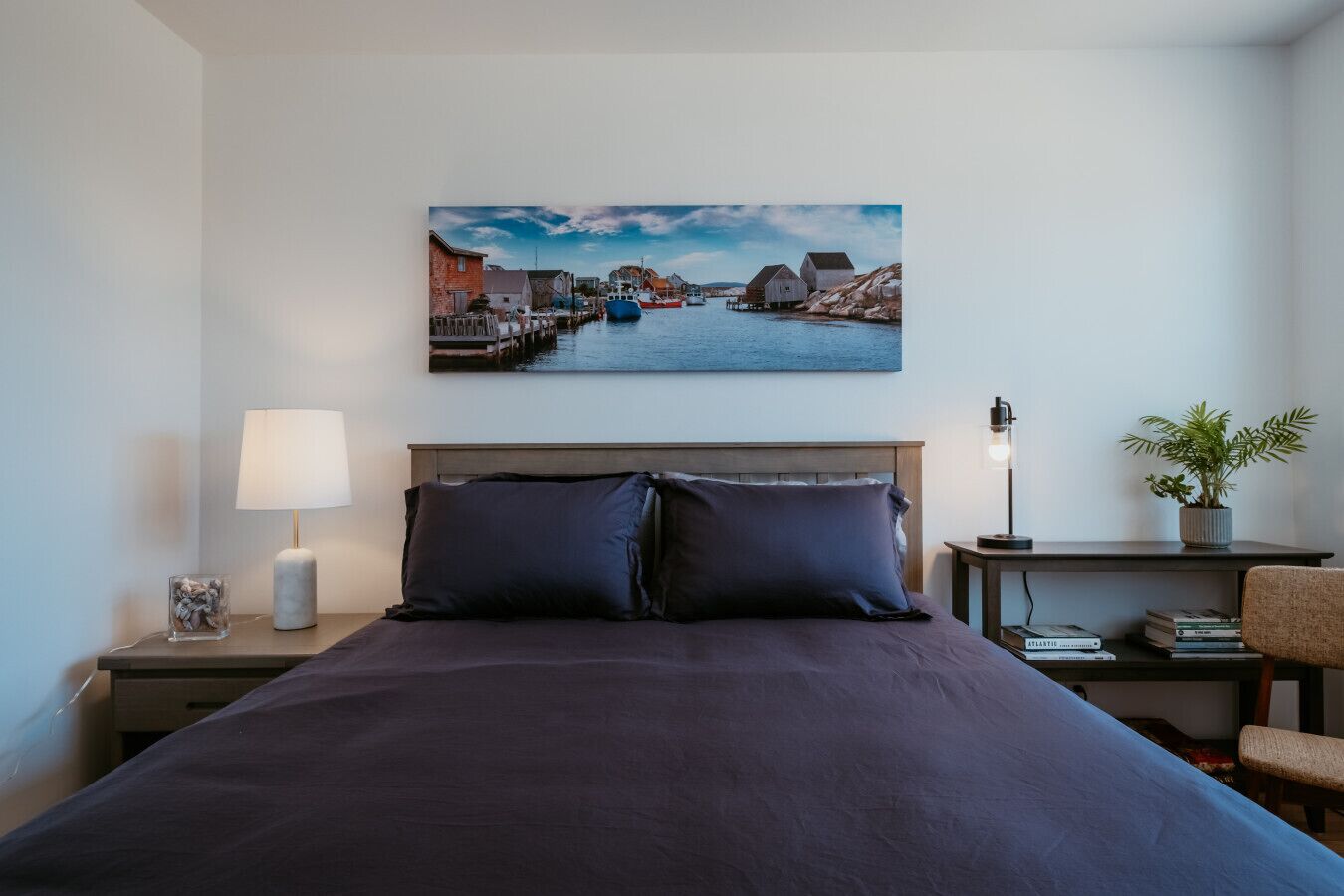 A hotel room with a bed, a nightstand with a lamp, a small table with a plant, and a wall-mounted painting of a coastal scene.