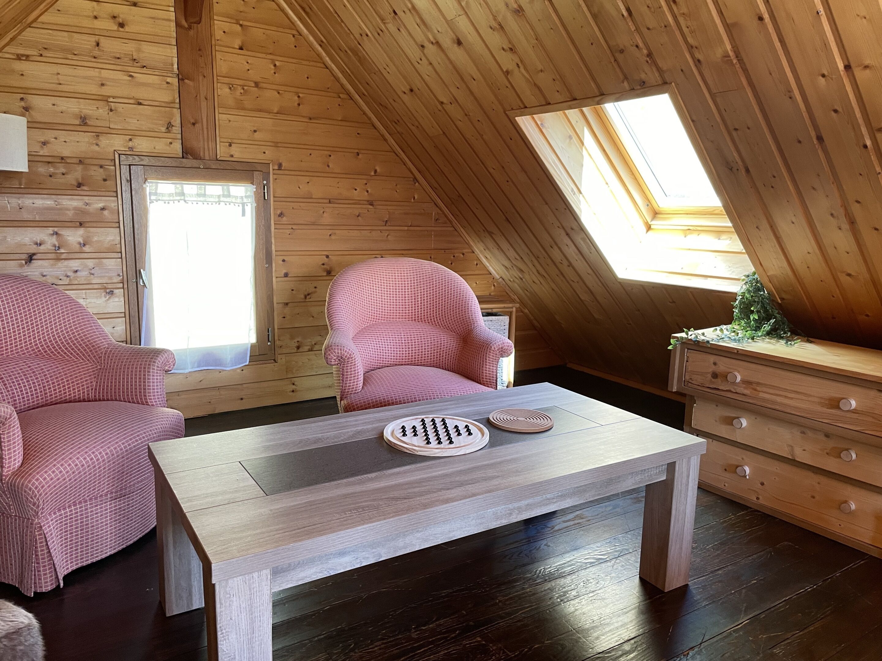 Una habitación acogedora con paredes de madera y techo inclinado, que incluye un sillón a cuadros, una mesita con un plato decorativo y un aparador de madera.