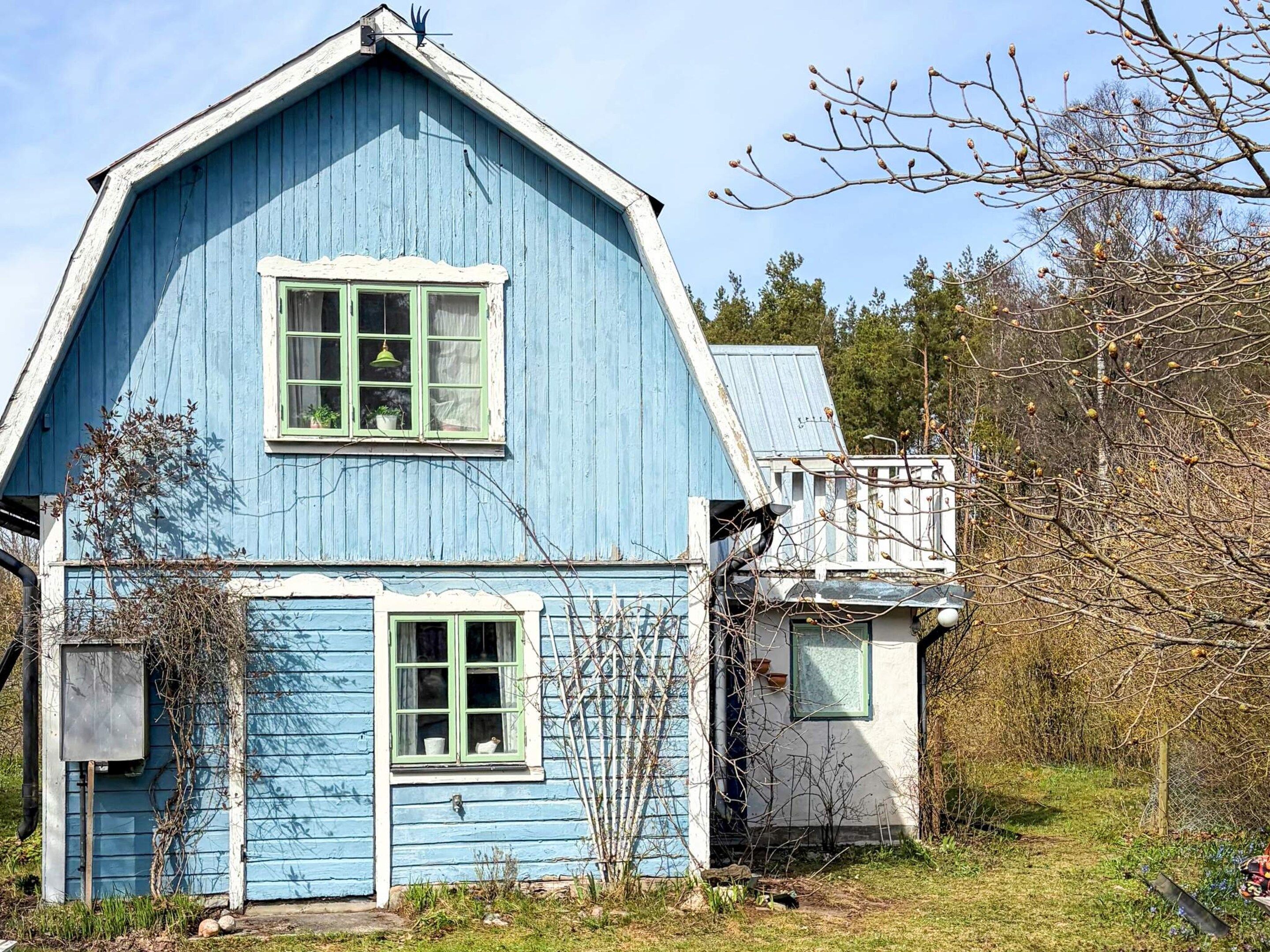 A blue wooden house with two windows, a porch, and a small attached structure.