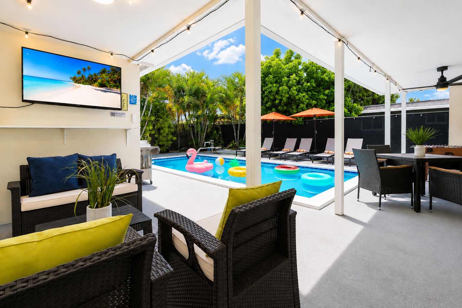 Un área de piscina al aire libre con sillones de descanso, una mesa de comedor y una pantalla de televisión que muestra una escena de playa.
