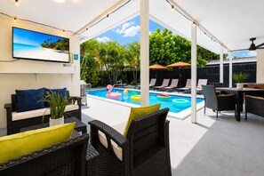 An outdoor pool area with lounge chairs, a dining table, and a television screen displaying a beach scene.