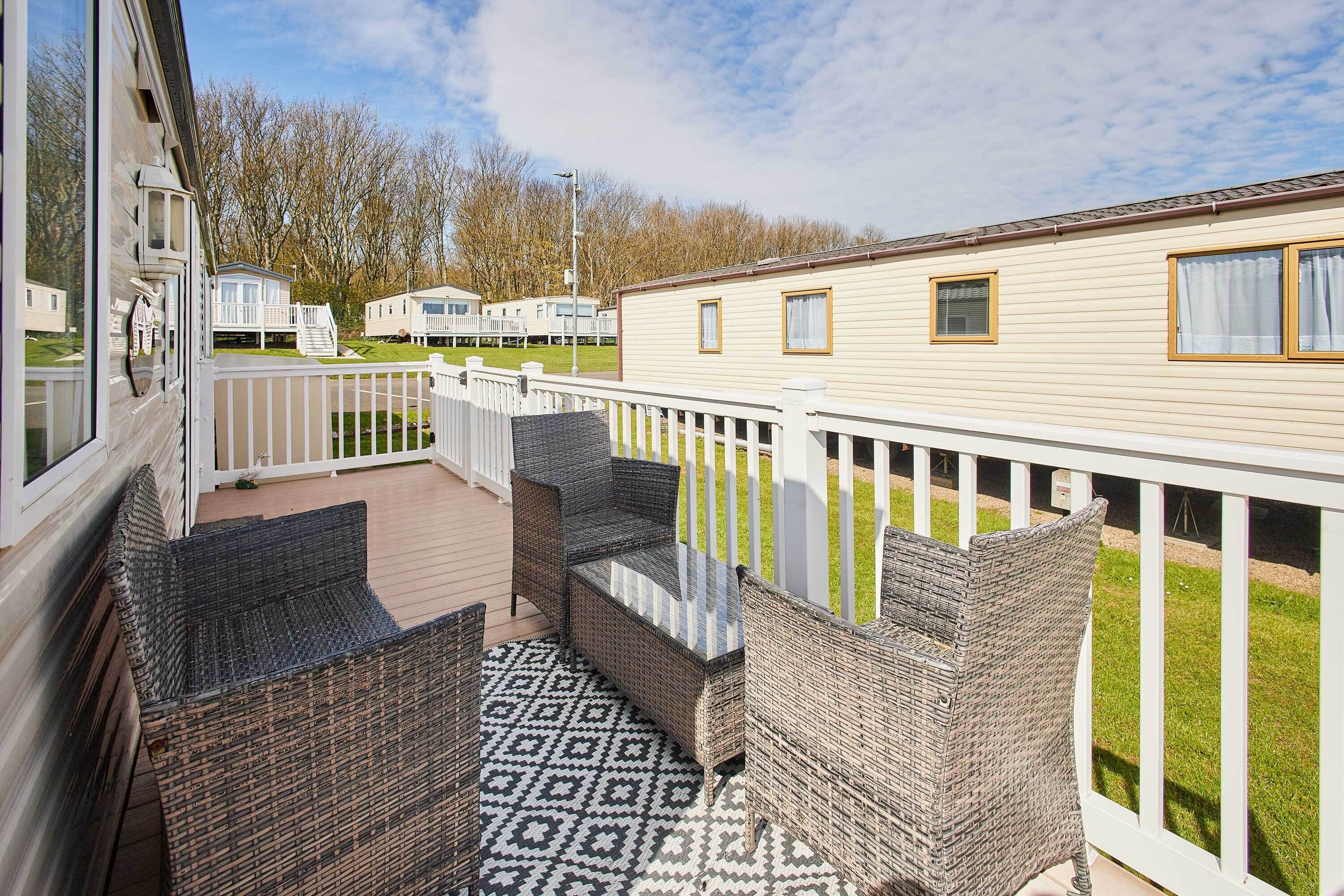A deck with wicker furniture and a patterned rug, surrounded by a white fence and a grassy area.