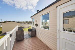 A mobile home with a deck, two wicker chairs, a small table, and a window with a view of other homes.