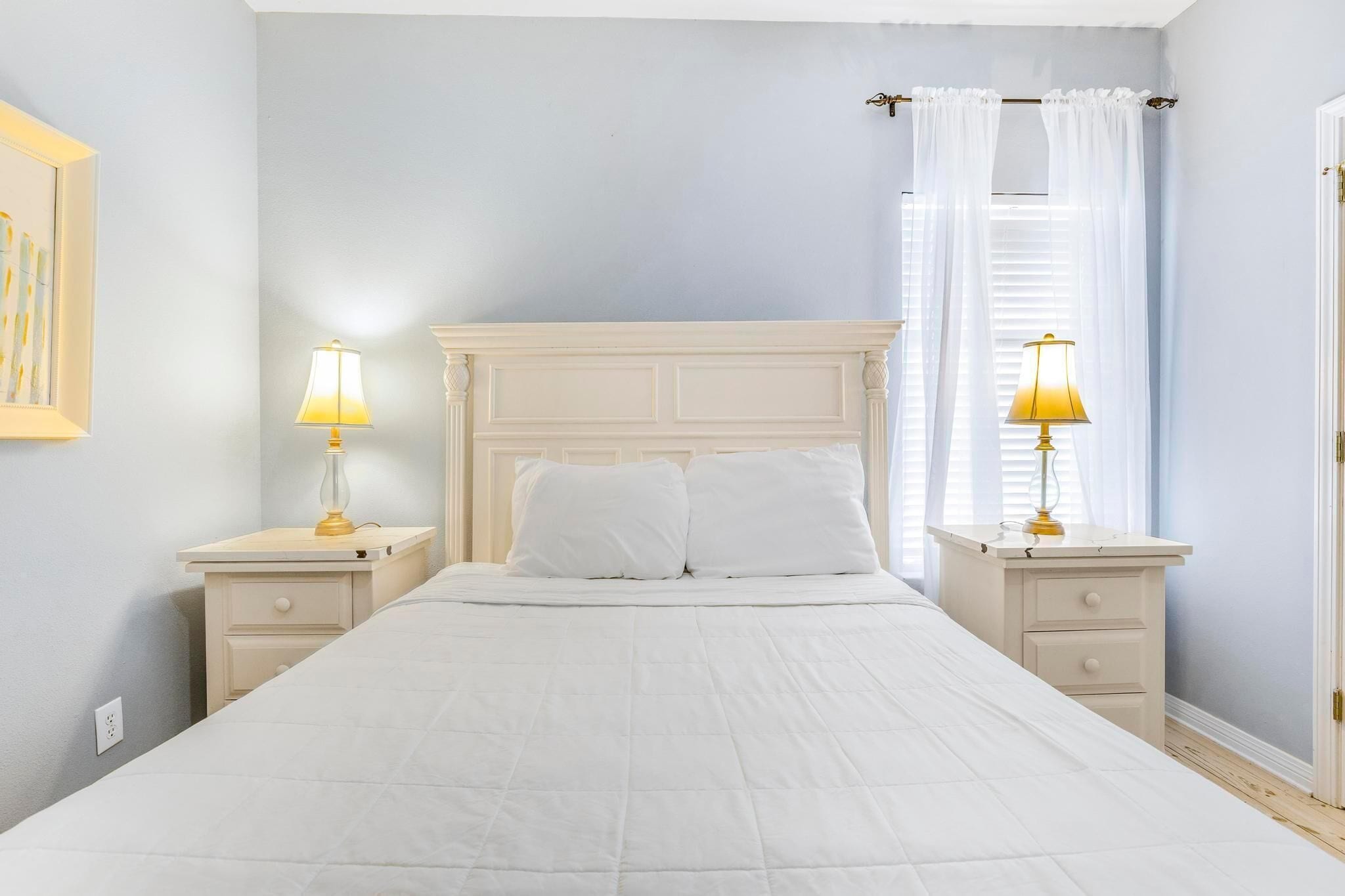 A bedroom with a white bed, two bedside tables with lamps, a white headboard, and a window with curtains.