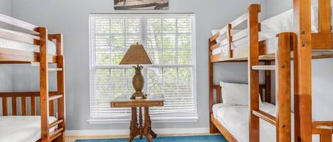 Une chambre avec deux lits superposés, une petite table avec une lampe, une fenêtre avec des stores et un tableau au mur.