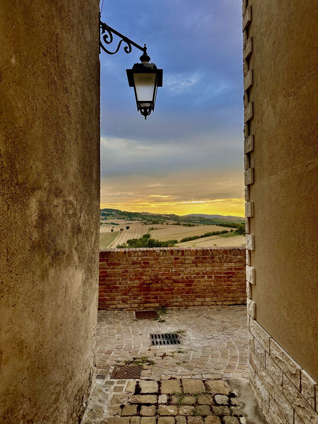 A stone-paved courtyard with a brick wall, a metal lantern, and a view of rolling hills under a clear sky.