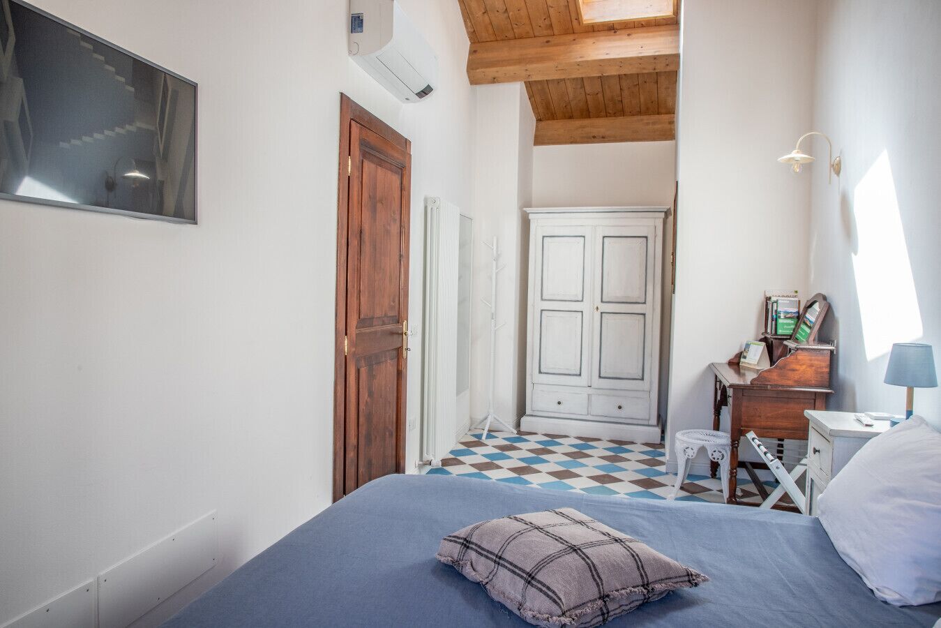A bedroom with a bed, a wooden door, a white wardrobe, a radiator, a desk with a lamp, and a checkered floor.