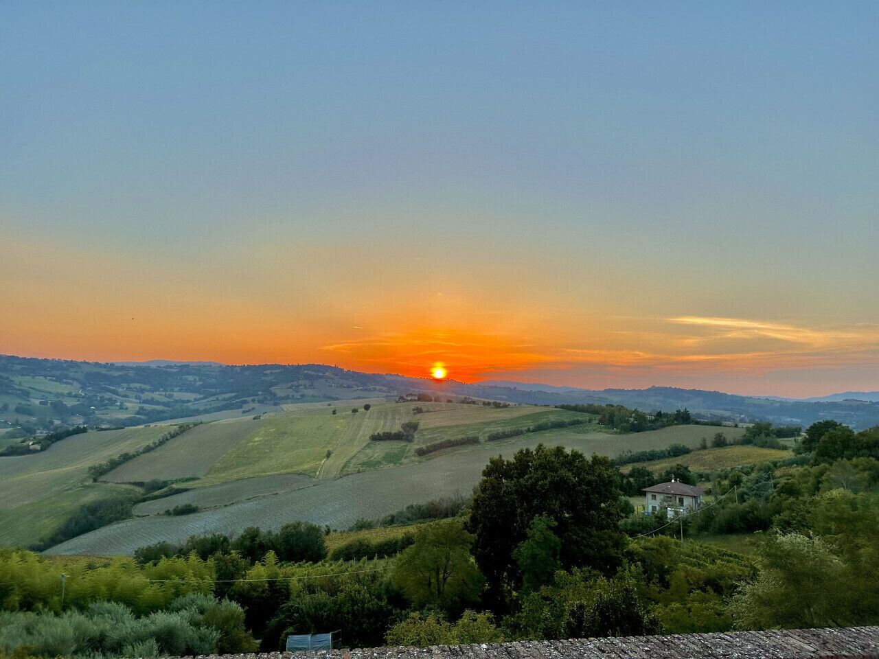 A rural landscape with rolling hills, a sunset, and a house in the foreground.
