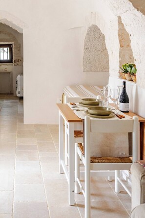 Uma área de refeições com uma mesa comprida posta para quatro pessoas, cadeiras brancas e uma parede de pedra rústica.