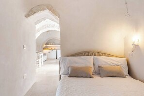 Quarto com cama, travesseiros, mesa de cabeceira, luminária na parede e uma porta arqueada que dá para uma cozinha.