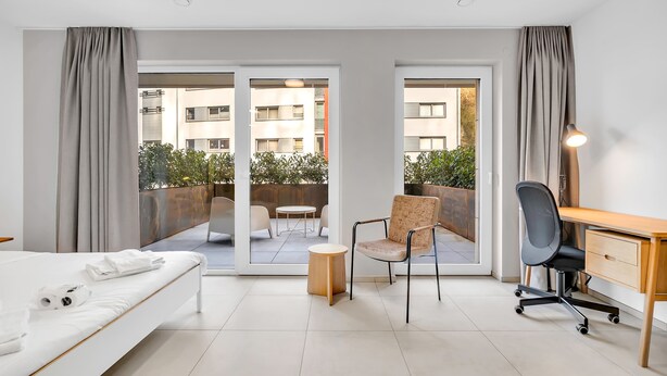 A hotel room with a bed, a desk, a chair, a small table, and a view of the outdoor balcony. - Neudorf Studio Near Kirchberg and Airport (Luxembourg)
