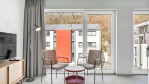 A modern room with a TV, two chairs, a small table, and a large window with a view of buildings and trees. - Modern Studio in Neudorf Near Kirchberg (Luxembourg)