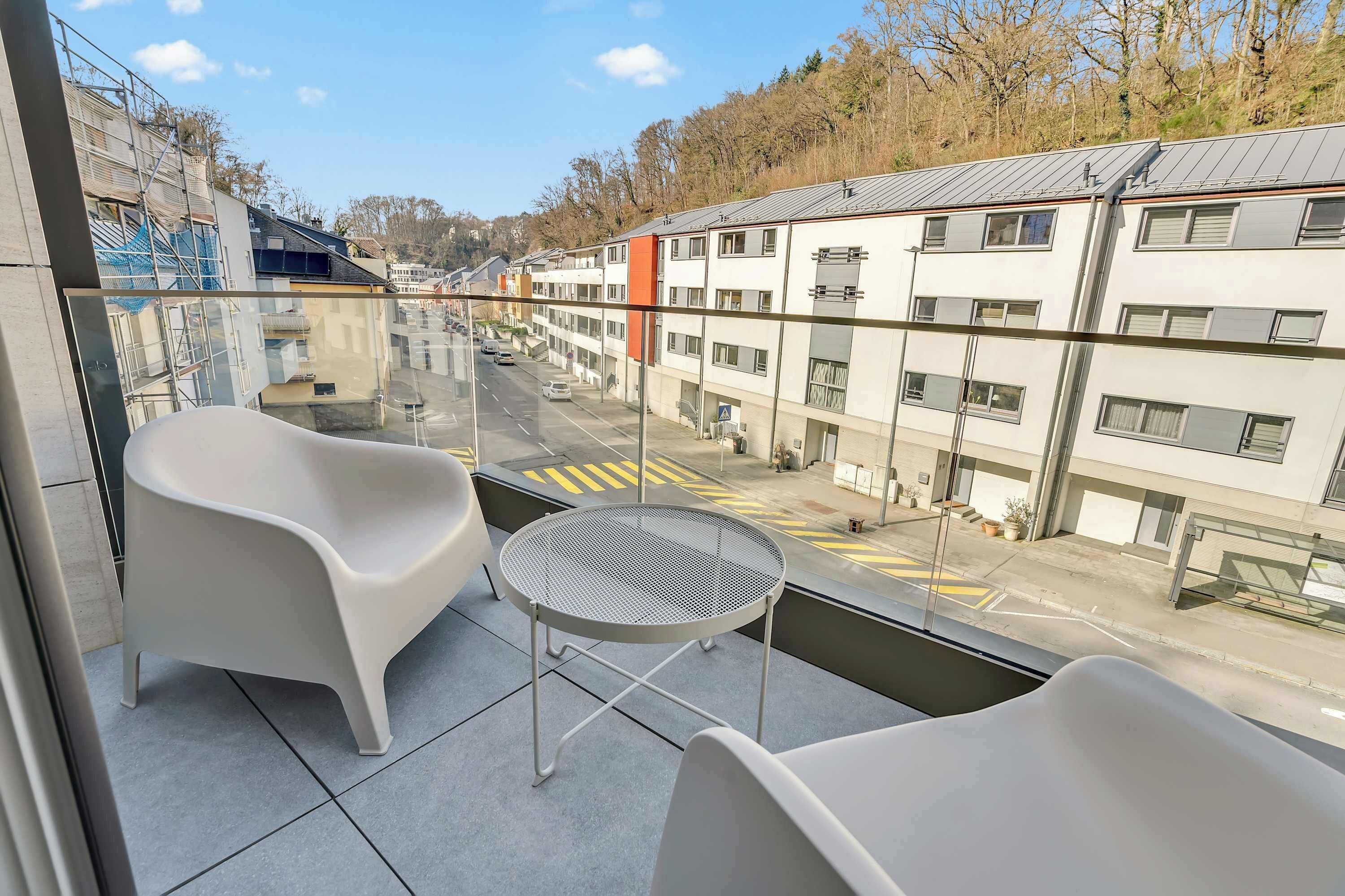 A balcony with two white chairs and a small round table, overlooking a street with buildings and a clear sky.