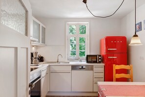 Cocina pequeña con muebles blancos, un refrigerador rojo, una silla de madera y una ventana con vistas a la vegetación.