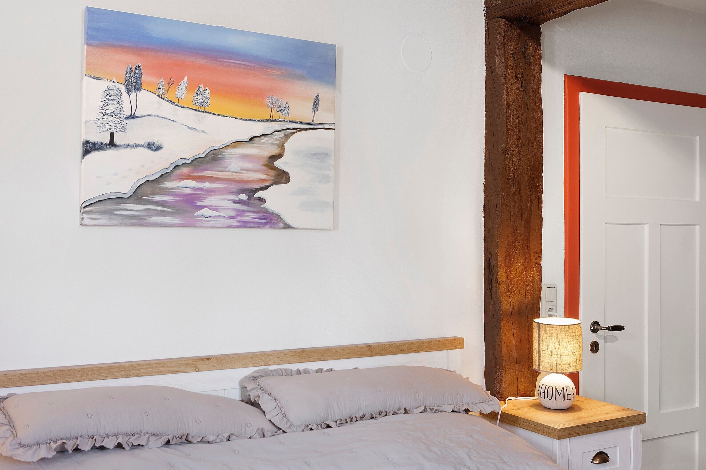 A bedroom with a bed, pillows, a bedside table with a lamp, a painting on the wall, and a door with a red frame.