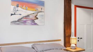 A bedroom with a bed, pillows, a bedside table with a lamp, a painting on the wall, and a door with a red frame.