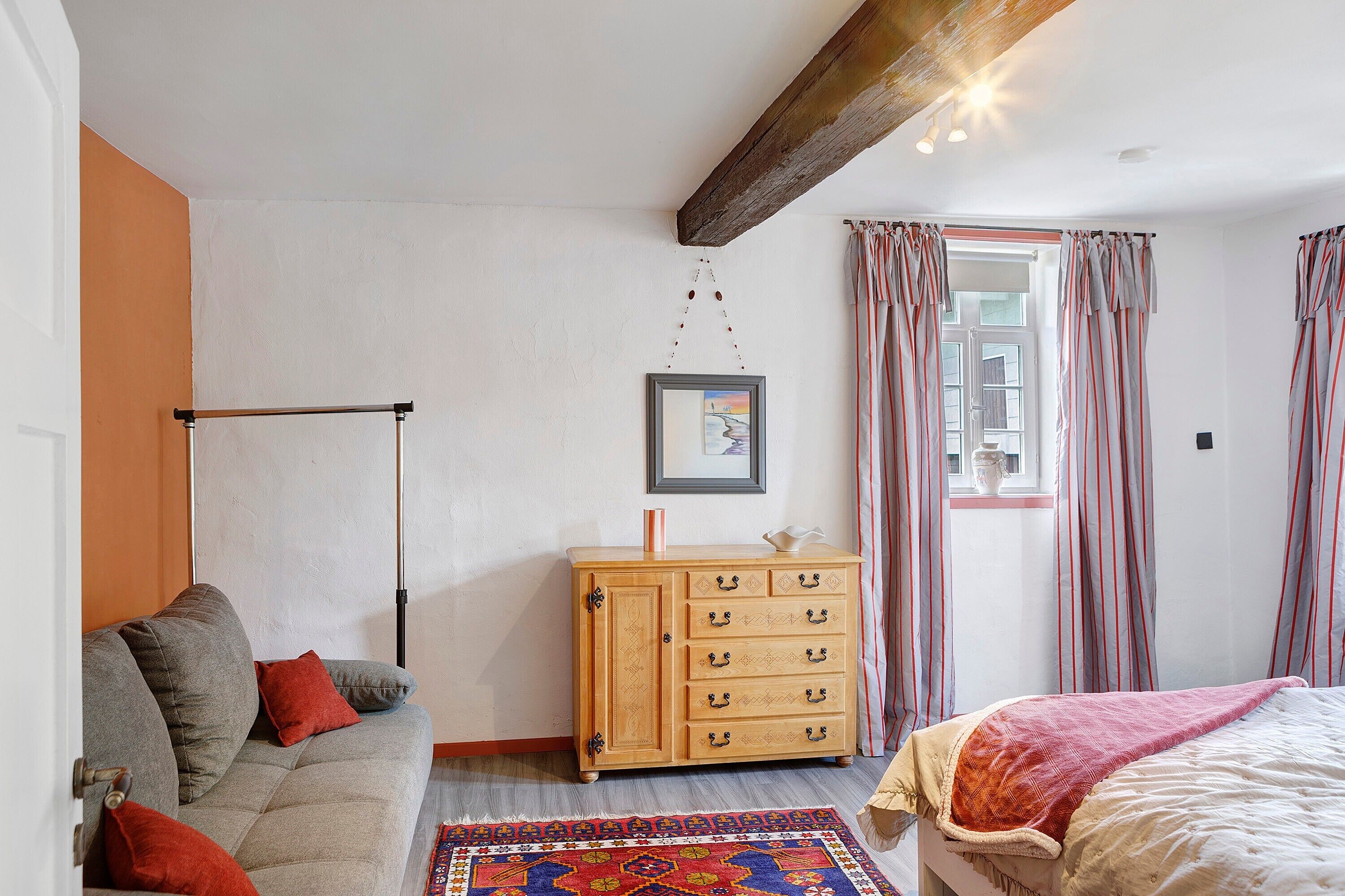 A bedroom with a sofa, a wooden dresser, a bed, a window with curtains, and a hanging light fixture.