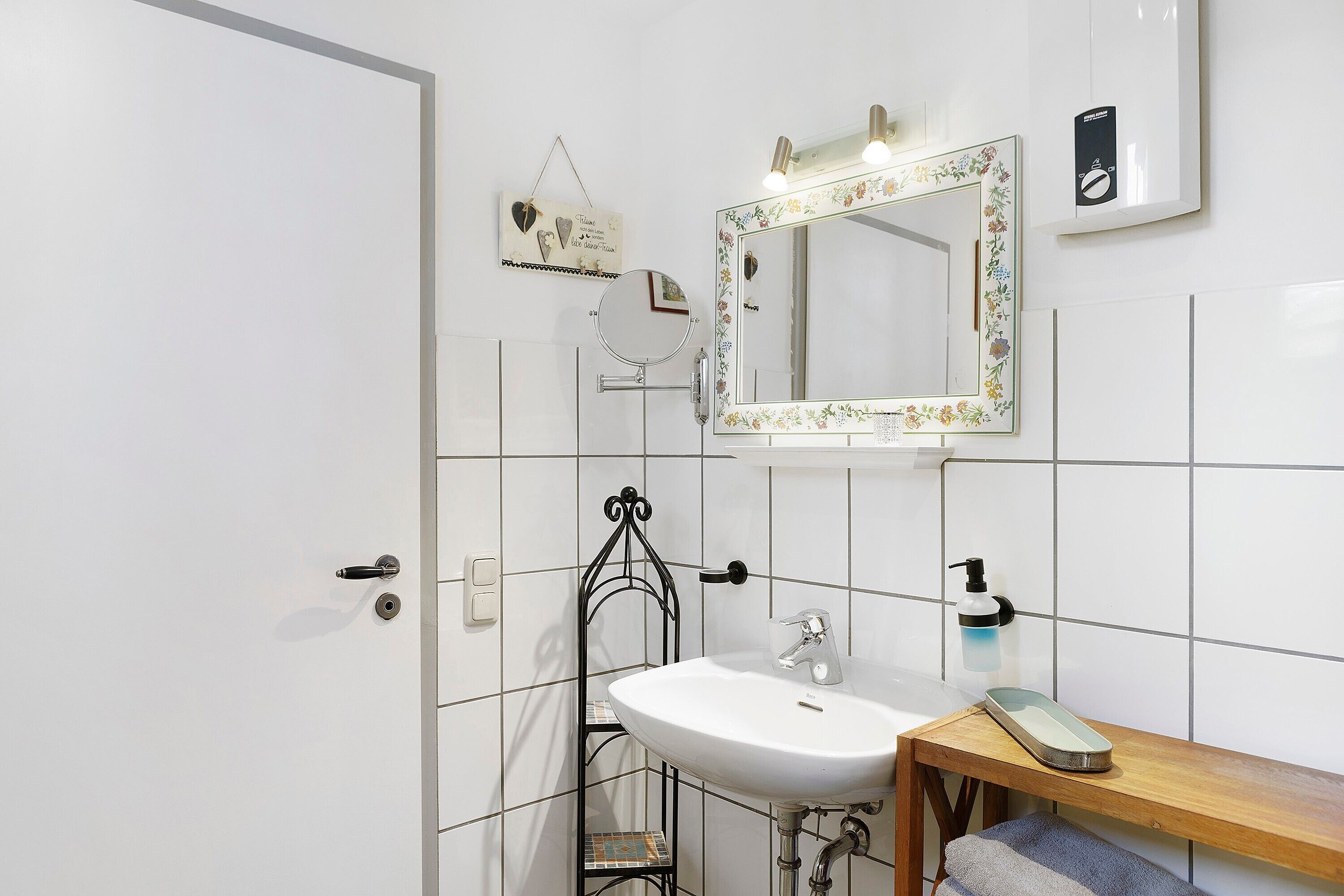 Un baño con lavamanos blanco, un espejo con marco floral, una pequeña estantería y un perchero para toallas.