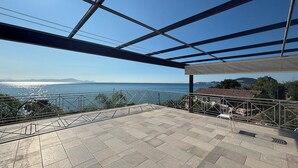 A terrace with a view of the sea and distant mountains, metal railings, and a tiled floor.
