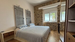 A bedroom with a stone wall, a bed with a white headboard, a nightstand, a wooden dresser, and a window with a view.
