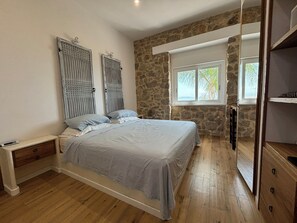 A bedroom with a stone wall, a bed with a white headboard, a nightstand, a wooden dresser, and a window with a view.