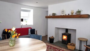 Een woonkamer met een open haard, een ronde houten tafel, een rode bank en een raam met jaloezieën.