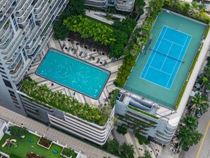 Vista aérea de un edificio moderno con piscina en la azotea, cancha de tenis y vegetación circundante.