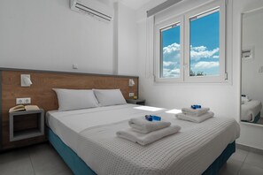 Habitación de hotel con cabecera de madera, una cama grande con ropa blanca, una mesita de noche con un teléfono y un libro, una ventana con vistas al cielo y un espejo que refleja la cama.