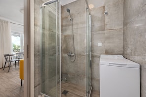 A modern bathroom with a glass-enclosed shower, a white washing machine, and a dining area with a yellow chair. - SLV Danai 7 Apartments (Pangaio)