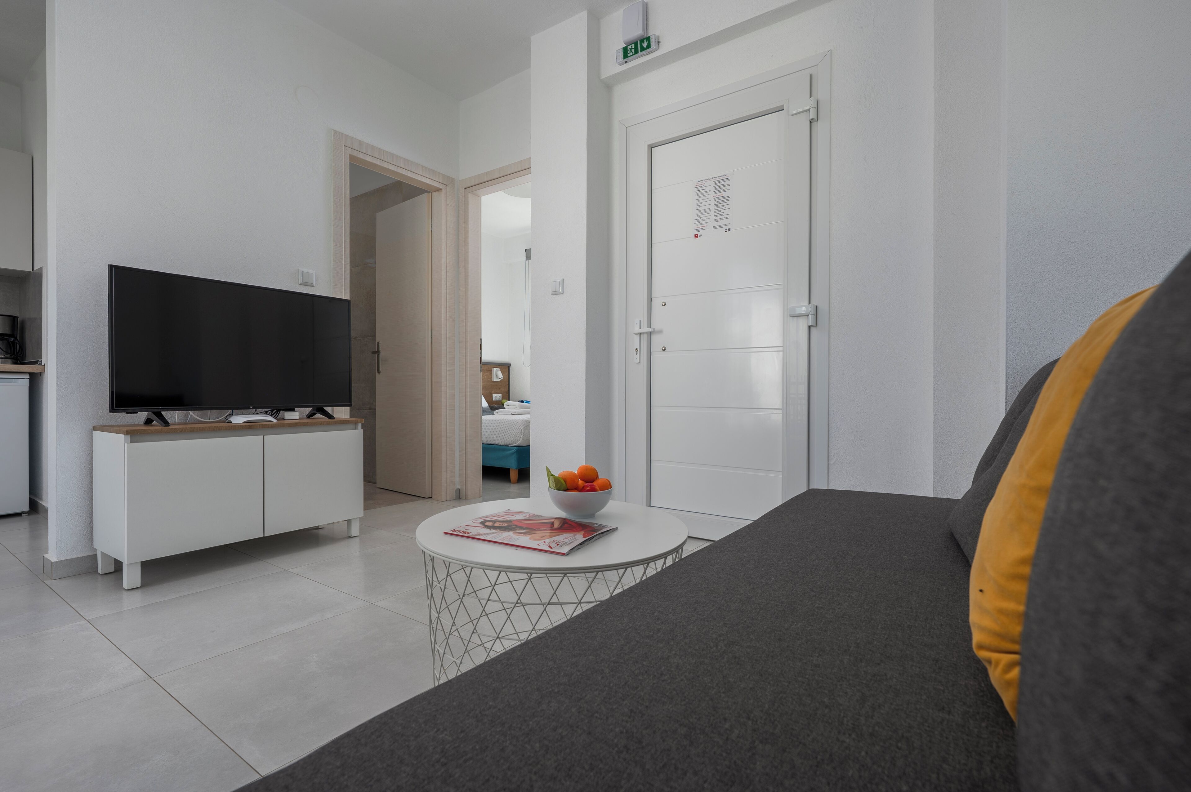 Un salon moderne avec un téléviseur à écran plat, une console blanche, une table basse ronde sur laquelle se trouve un saladier rempli de fruits, et un canapé agrémenté d’un coussin.