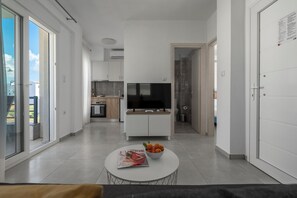 A modern living room with a flat-screen TV, a round coffee table with a magazine and fruit bowl, and a sliding glass door leading to a balcony. - SLV Danai 7 Apartments (Pangaio)