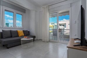 Una sala moderna con un sofá gris, una mesita redonda con fruta, un televisor y un balcón con puerta de vidrio.