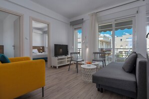 Ein modernes Wohnzimmer mit einer Couch, einem gelben Sessel, einem Fernseher, einem Esstisch und einem Balkon mit Blick auf Gebäude.