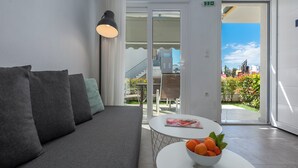 Ein modernes Hotelzimmer mit grauem Sofa, einem weißen Tisch mit Obstschale und Blick ins Freie.