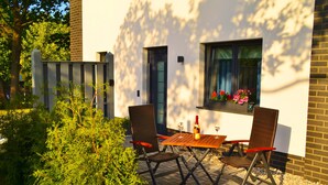 Een terras buiten met een tafel, stoelen, een fles wijn en een raam vol bloemen.