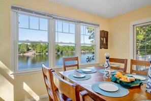Comedor con una mesa preparada para cuatro personas, ventanas amplias con vista a un lago y una silla de madera.