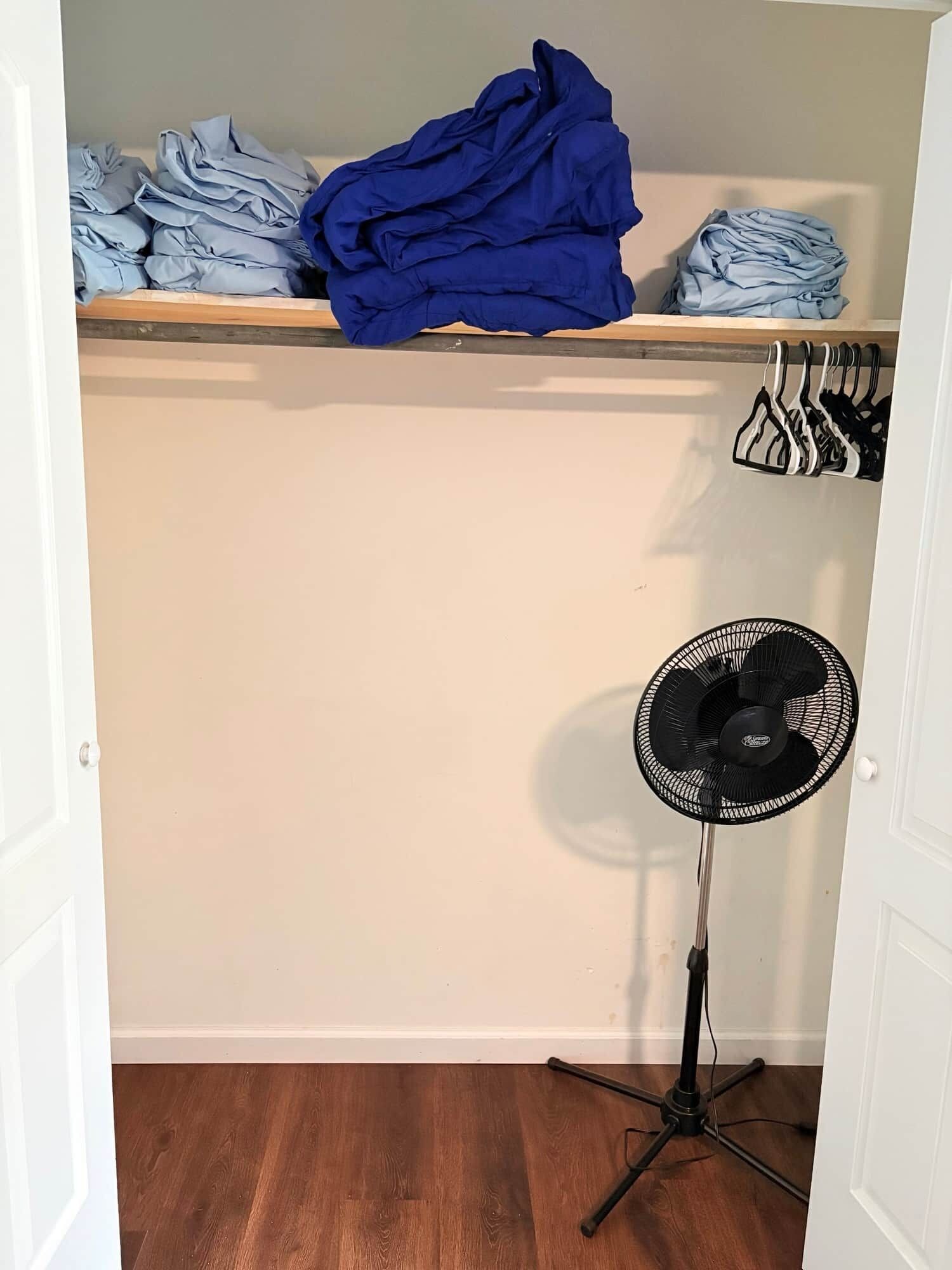 A room with a wooden floor, a standing fan, and a shelf with folded clothes.