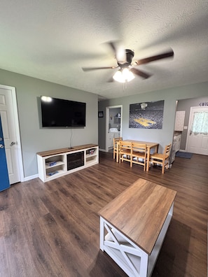 A living room with a TV, wooden furniture, a ceiling fan, and a sports poster.