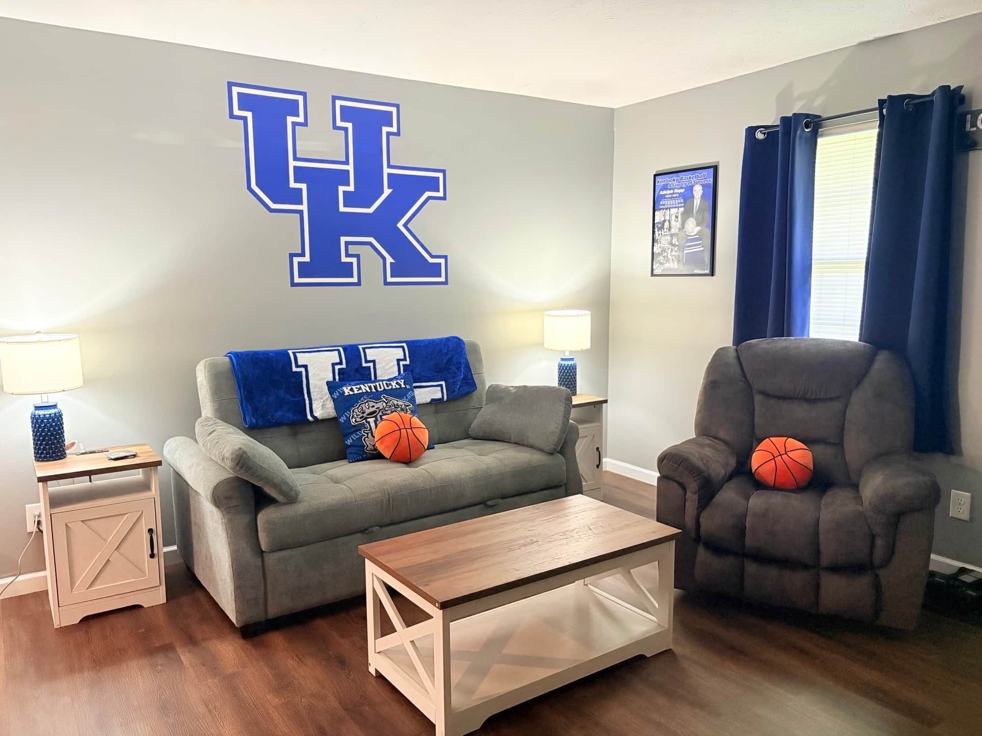 Un salon avec un grand logo « UK » sur le mur, un canapé gris, un fauteuil marron, une table basse et une petite table d’appoint avec une lampe.
