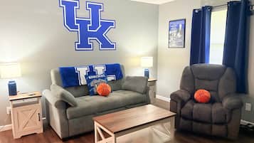 Un salon avec un grand logo « UK » sur le mur, un canapé gris, un fauteuil marron, une table basse et une petite table d’appoint avec une lampe.