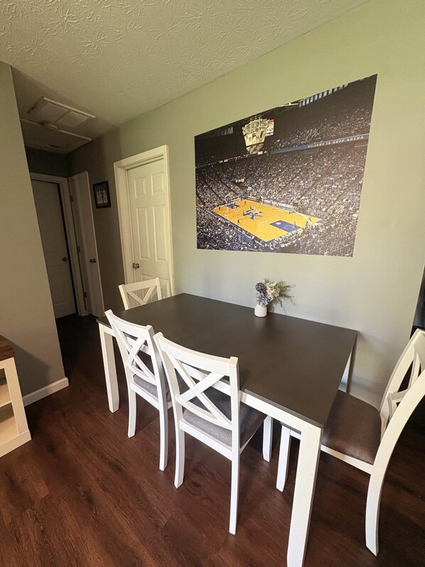 A dining area with a dark wooden table, white chairs, and a basketball court poster on the wall. - Ruppside Retreat I Cozy 2BR I 15m to RRG I Wi-Fi (Stanton)