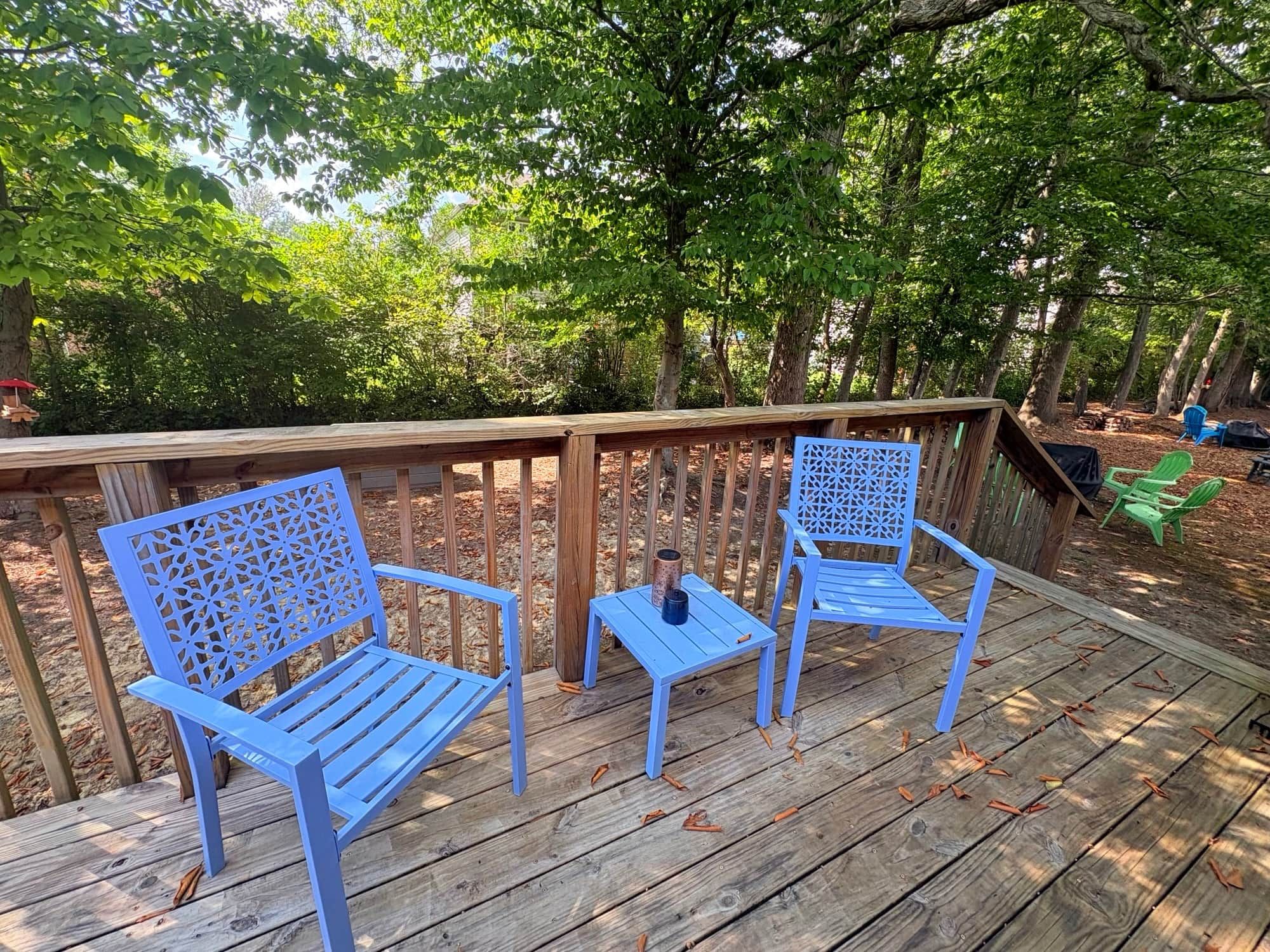 A wooden deck with two blue chairs and a small table, surrounded by trees.