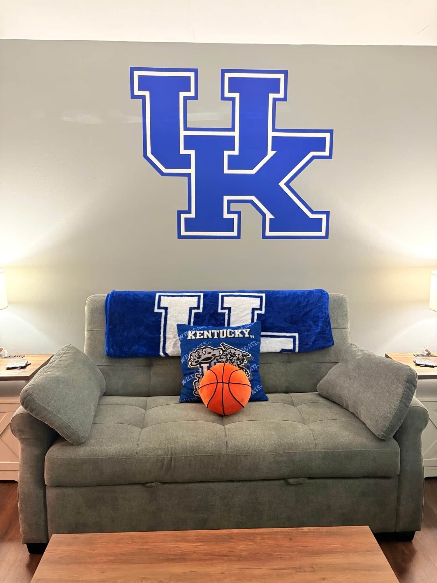 Un canapé gris sur lequel est posée une couverture bleue et des coussins ornés du logo du Royaume-Uni et du mot « KENTUCKY ». Un ballon de basket est placé sur le canapé.
