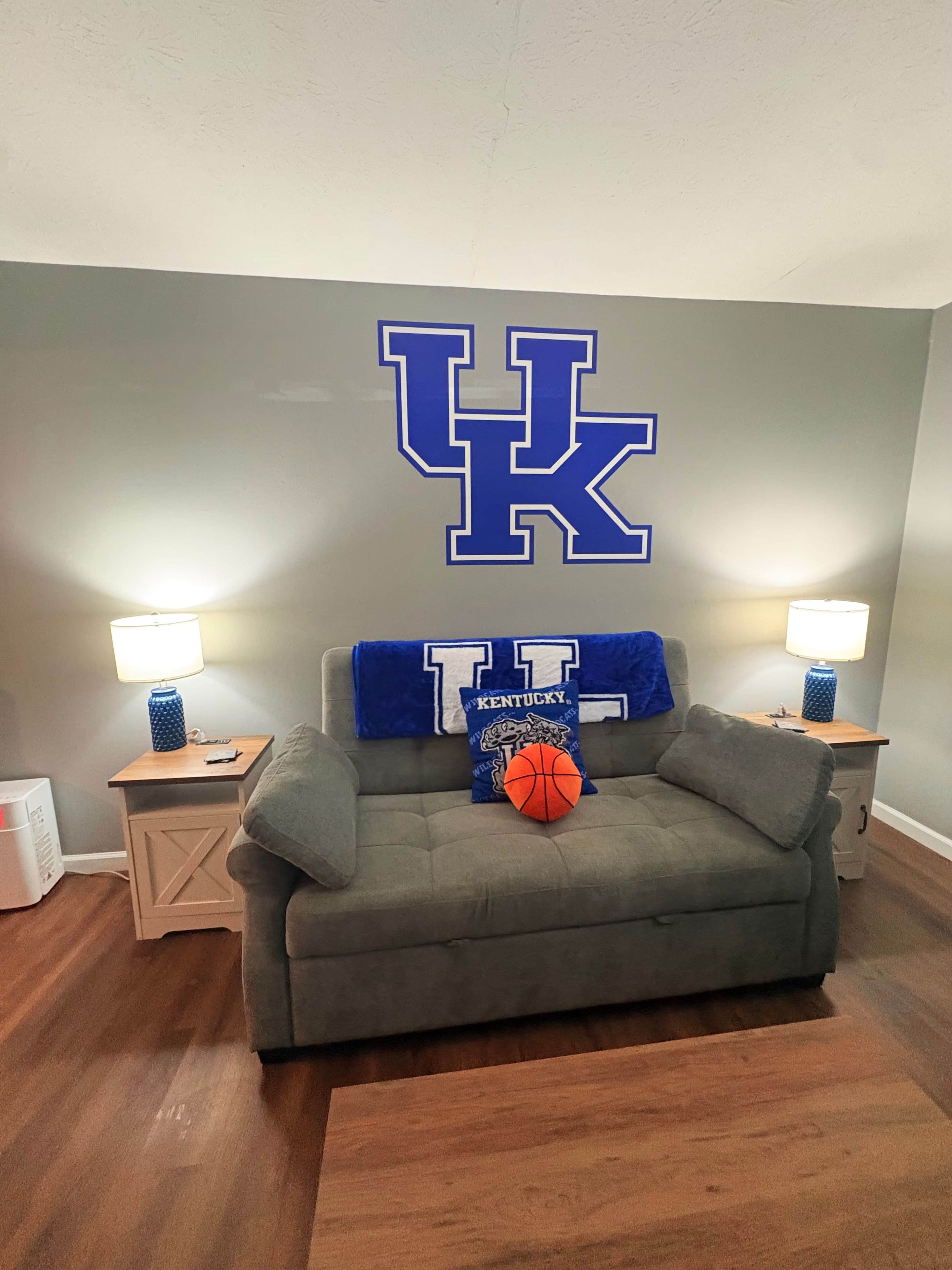 Ein Zimmer mit einem grauen Sofa, zwei Nachttischen mit Lampen, einer großen Wanddekoration mit den Buchstaben 'UK' und einem Basketball auf dem Sofa.